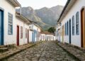 Rua íngreme com calçamento irregular de pedra pé de moleque, ladeada por casarios coloniais de fachada branca e esquadrias de madeira pintadas em tons vibrantes de azul, vermelho e amarelo. Ao fundo, ergue-se uma imponente formação montanhosa de pedra sob um céu claro.