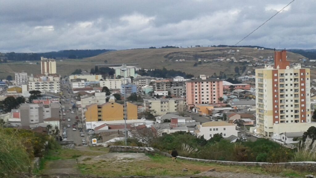 Visão ampla da área urbana de São Joaquim, mostrando prédios de diferentes alturas e casas espalhadas pelas colinas da região serrana. O céu está encoberto por nuvens densas, destacando o clima frio da cidade.