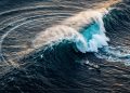 Onda gigante no oceano vista do alto, com navio cargueiro próximo e mar agitado em cenário de risco marítimo.