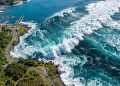 Vista aérea de ondas gigantes atingindo região costeira, com estrada, porto pequeno e vegetação próxima ao mar.