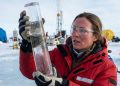 Cientista em ambiente polar segura um cilindro de gelo para análise, usando casaco vermelho, luvas e óculos de proteção em campo de pesquisa.