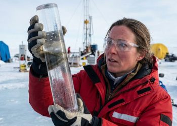 Cientista em ambiente polar segura um cilindro de gelo para análise, usando casaco vermelho, luvas e óculos de proteção em campo de pesquisa.