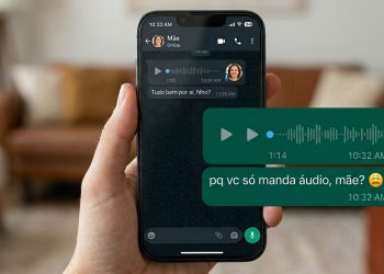Uma mão segurando um iPhone com a tela exibindo um chat do WhatsApp no modo escuro com a "Mãe". A conversa contém mensagens de áudio trocadas entre os dois e um texto do filho perguntando: "pq vc só manda áudio, mãe?" acompanhado de emojis de cansaço e dúvida. Uma sala de estar aparece desfocada ao fundo.