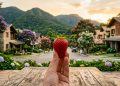 Mão segura morango maduro em frente a mesa de madeira, com casas, jardins floridos e montanhas ao fundo ao entardecer.