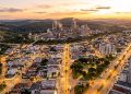 Vista aérea de Votorantim ao entardecer, com avenidas iluminadas, bairros urbanos, áreas verdes, morros e complexo industrial ao fundo.