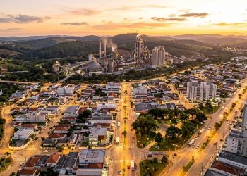 Vista aérea de Votorantim ao entardecer, com avenidas iluminadas, bairros urbanos, áreas verdes, morros e complexo industrial ao fundo.