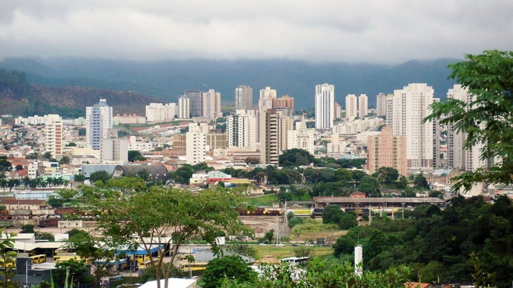 Vista panorâmica da zona urbana de Jundiaí, com prédios altos, áreas verdes, montanhas e céu nublado ao fundo.