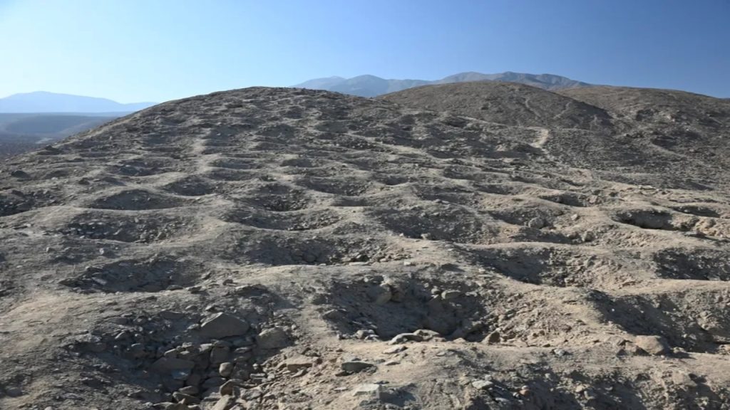 Paisagem árida e rochosa com colinas, solo seco, formações irregulares e montanhas ao fundo sob céu azul.