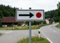 O que significa a placa de trânsito branca com uma seta preta apontando para a direita e um pequeno círculo vermelho?